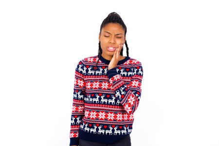 Foto de Concepto de dolor de muelas. Indoor shot of young African American woman wearing Christmas sweater against white wall feeling pain, holding his cheek with hand, suffering of bad dothache, looking at camera with painful expression - Imagen libre de derechos