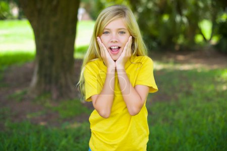 Foto de Molesto niña caucásica usando camiseta amarilla de pie al aire libre tocando la cara con dos manos - Imagen libre de derechos