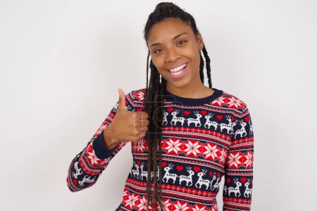 Foto de Mujer afroamericana vistiendo suéter de Navidad contra la pared blanca dando el pulgar hacia arriba gesto, buen trabajo! Emoción humana positiva expresión facial lenguaje corporal. - Imagen libre de derechos