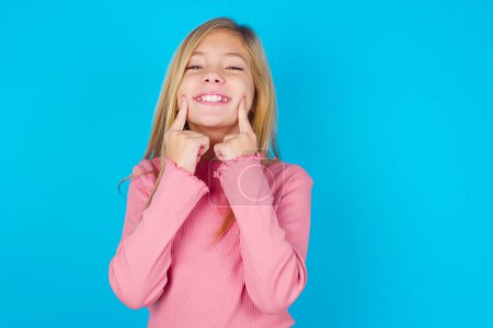 Photo for Happy pretty teen girl with toothy smile, keeps index fingers near mouth, fingers pointing and forcing cheerful smile - Royalty Free Image
