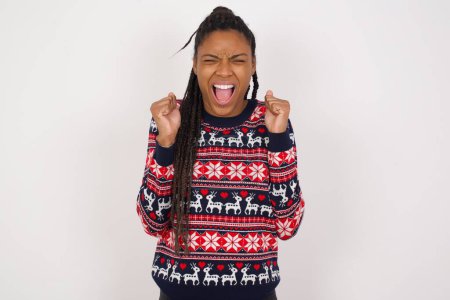 Photo for African American woman wearing Christmas sweater against white wall rejoicing his success and victory clenching fists with joy being happy to achieve aim and goals. Positive emotions, feelings. - Royalty Free Image
