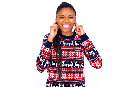 Téléchargez les photos : Arrête de faire ce bruit ennuyeux ! Malheureux stressé femme afro-américaine portant un pull de Noël contre le mur blanc faisant face à l'inquiétude, bouchant les oreilles avec les doigts, irrité par un bruit fort. - en image libre de droit