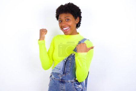 Foto de Hurra fresca joven mujer afroamericana con el pelo corto con denim en general contra blanco punta de la pared detrás de puño de mano espacio vacío - Imagen libre de derechos