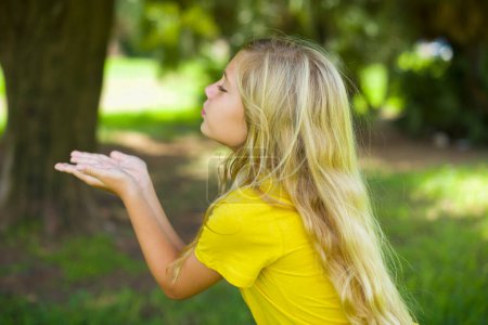 Photo for Profile side view view portrait of attractive caucasian little girl wearing yellow t-shirt standing outdoors sending air kiss - Royalty Free Image