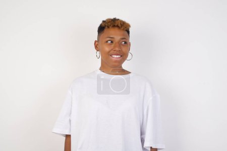 Téléchargez les photos : Dreamy jeune femme afro-américaine portant un t-shirt blanc avec une expression réfléchie, regarde loin garde les mains vers le bas en mordant sa lèvre pense à quelque chose d'agréable pose contre le mur gris. - en image libre de droit