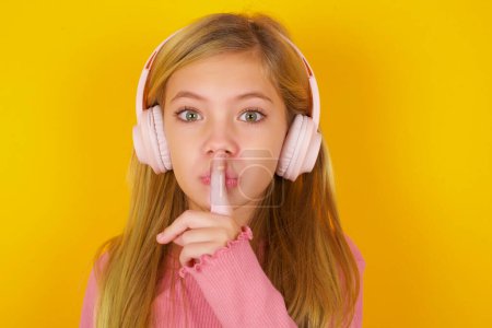 Photo for Pretty teen girl making hush gesture with finger on her lips wearing  wireless headphones. Be quiet. - Royalty Free Image