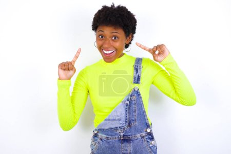 Photo for Cheerful young African American woman with short hair wearing denim overall against white wall demonstrating hairdo - Royalty Free Image
