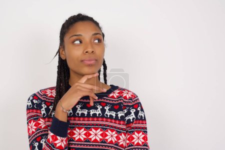 Photo for Dreamy African American woman wearing Christmas sweater against white wall with pleasant expression, looks sideways, keeps hand under chin, thinks about something pleasant. - Royalty Free Image