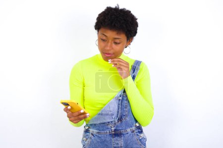 Foto de Pensativo hembra afroamericana feliz con rizado tupida usa pantalones vaqueros sobre blanco pared mantenga mirada teléfono, copyspace - Imagen libre de derechos