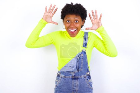 Foto de Joven mujer afroamericana con el pelo corto con denim en general contra la pared blanca se ve con emoción en la cámara, mantiene las manos levantadas sobre la cabeza, se da cuenta de algo inesperado, reacciona ante noticias repentinas. - Imagen libre de derechos
