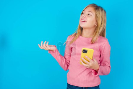 Happy pleased pretty teen girl raises palm and holds cellphone uses high speed internet for text messaging or video calls