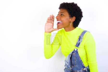 Foto de Joven mujer afroamericana con el pelo corto con denim en general contra la pared blanca mirada espacio vacío sosteniendo la mano cerca de su cara y gritando o llamando a alguien. - Imagen libre de derechos