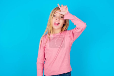 Photo for Pretty teen girl gestures with finger on forehead makes loser gesture makes fun of people shows tongue - Royalty Free Image