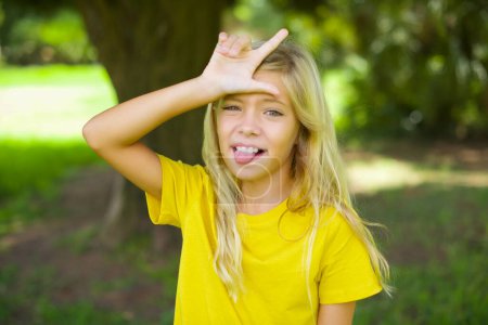 Foto de Divertido caucásico niña usando amarillo camiseta de pie al aire libre hace gesto perdedor burlándose de alguien sobresale lengua haciendo mueca cara. - Imagen libre de derechos