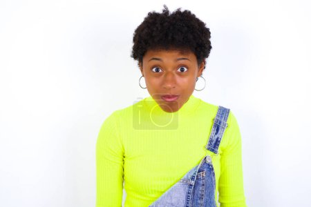 Foto de Joven mujer afroamericana aturdida con el pelo corto usando denim en general contra las miradas de la pared blanca reacciona ante noticias impactantes. MODELO Asombroso sostiene la respiración - Imagen libre de derechos