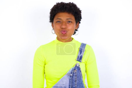 young African American woman with short hair wearing denim overall against white wall puffing cheeks with funny face. Mouth inflated with air, crazy expression.