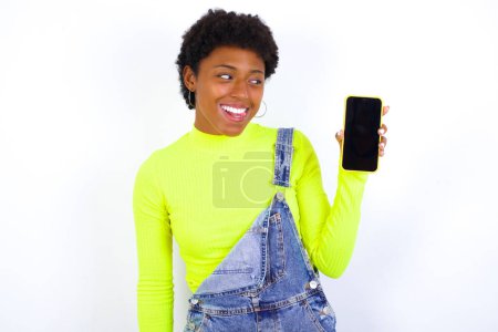 Photo for Cheerful cheery content African American female with curly bushy wears jeans overalls over white wall holding in hands device hobby smm post blog - Royalty Free Image