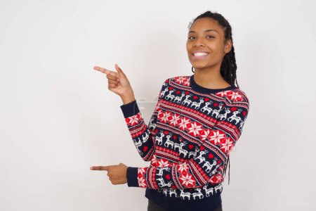 Téléchargez les photos : Femme afro-américaine portant un pull de Noël contre les points de mur blanc de côté avec une expression surprise avec la bouche ouverte, montre quelque chose d'étonnant. Concept de publicité. - en image libre de droit