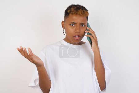 Téléchargez les photos : Jeune femme afro-américaine portant un t-shirt blanc parlant au téléphone sur fond isolé stressé avec la main sur le visage, choqué par la honte et le visage surprise, en colère et frustré. Peur et contrariété par erreur. - en image libre de droit