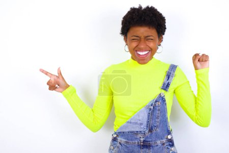 Foto de Joven mujer afroamericana alegre con el pelo corto que usa el denim en general contra la pared blanca que muestra el anuncio del espacio de copia que celebra suerte - Imagen libre de derechos