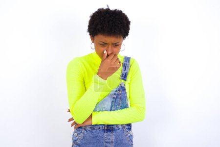 Téléchargez les photos : Jeune afro-américaine aux cheveux courts portant globalement du denim contre du blanc tenant son nez à cause d'une mauvaise odeur. - en image libre de droit