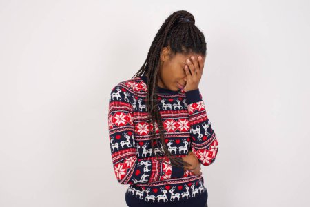 Foto de Mujer afroamericana usando suéter de Navidad contra la pared blanca haciendo gesto de facepalm mientras sonríe sorprendido con la situación estúpida. - Imagen libre de derechos