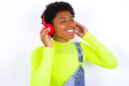Photo for Young African American woman with short hair wearing denim overall against white wall with headphones on her head, listens to music, enjoying favourite song with closed eyes, holding hands on headset. - Royalty Free Image