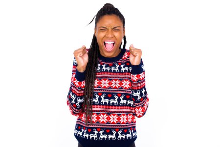 Téléchargez les photos : Femme afro-américaine portant un pull de Noël contre un mur blanc se réjouissant de son succès et de la victoire serrant les poings avec joie d'être heureux d'atteindre l'objectif et les objectifs. Émotions positives, sentiments. - en image libre de droit