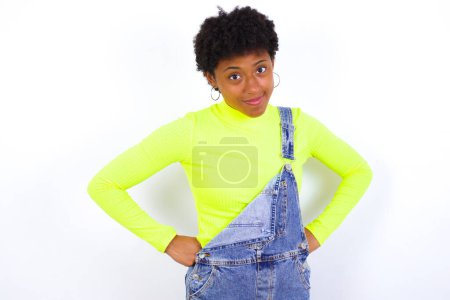 Photo for Funny frustrated young African American woman with short hair wearing denim overall against white wall holding hands on waist and silly looking at awkward situation. - Royalty Free Image