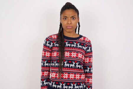 Téléchargez les photos : Femme afro-américaine portant un pull de Noël contre le mur blanc fronçant les sourcils étant mécontent de quelque chose. - en image libre de droit