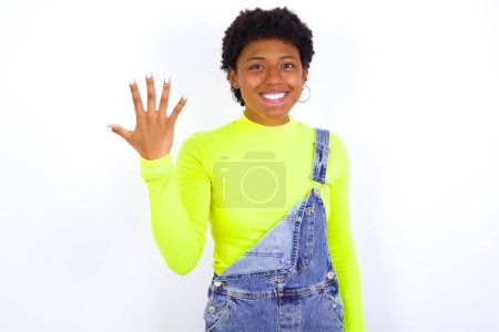Téléchargez les photos : Jeune femme afro-américaine avec les cheveux courts portant le denim dans l'ensemble contre le mur blanc souriant et regardant amical, montrant numéro cinq ou cinquième avec la main vers l'avant, compte à rebours - en image libre de droit