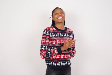 Foto de Mujer afroamericana vistiendo suéter de Navidad contra pared blanca sintiéndose feliz, sonriendo y aplaudiendo, diciendo felicitaciones con un aplauso. - Imagen libre de derechos