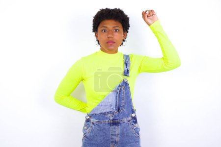 Foto de Joven mujer afroamericana con el pelo corto vistiendo denim en general contra la pared blanca sintiéndose seria, fuerte y rebelde, levantando el puño, protestando o luchando por la revolución. - Imagen libre de derechos