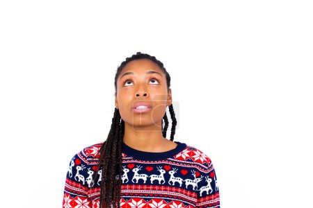 Téléchargez les photos : Femme afro-américaine portant un pull de Noël contre un mur blanc levant les yeux alors qu'il voit quelque chose d'étrange. - en image libre de droit