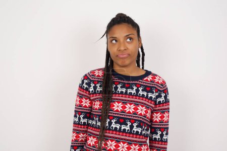 Foto de Mujer afroamericana vistiendo suéter de Navidad contra la pared blanca tiene la cara preocupada mirando los labios juntos, estar molesto pensando en algo importante, mantiene las manos abajo. - Imagen libre de derechos