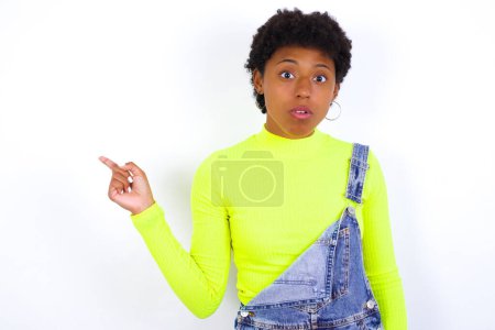 Foto de Emotiva mujer afroamericana joven con el pelo corto que lleva denim en general contra la pared blanca mantiene la mandíbula caída de choque demuestra puntos de promoción increíbles a la derecha en el espacio en blanco demuestra gran venta de compras. Publicidad - Imagen libre de derechos