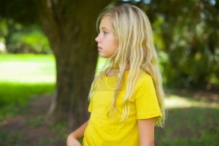 Téléchargez les photos : Petite fille caucasienne sérieuse portant un t-shirt jaune debout à l'extérieur garde les mains croisées se tient dans la pose réfléchie concentrée quelque part - en image libre de droit