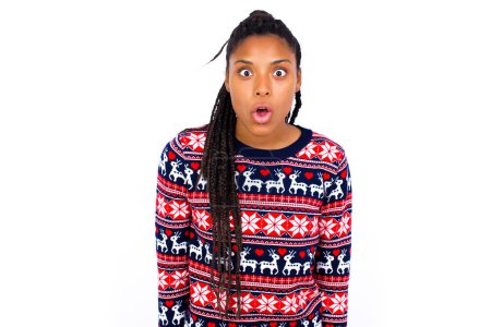 Téléchargez les photos : Femme afro-américaine portant un pull de Noël contre un mur blanc ayant regard stupéfait et choqué, avec la bouche ouverte et la mâchoire tombée en s'écriant : Wow, je ne peux pas croire cela. Surprise et choc - en image libre de droit