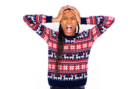 Foto de Pánico conmocionado Mujer afroamericana usando suéter de Navidad contra pared blanca cogida de la mano en la cabeza y gritando en desesperación y frustración. - Imagen libre de derechos