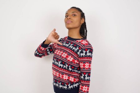 Photo for Closeup of cheerful African American woman wearing Christmas sweater against white wall looks joyful, satisfied and confident, points at himself with thumb. - Royalty Free Image