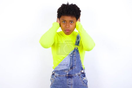 Foto de Mujer afroamericana frustrada joven con el pelo corto que usa denim en general contra la pared blanca tapar las orejas con las manos no quiere escuchar rock duro, ruido o música fuerte. - Imagen libre de derechos