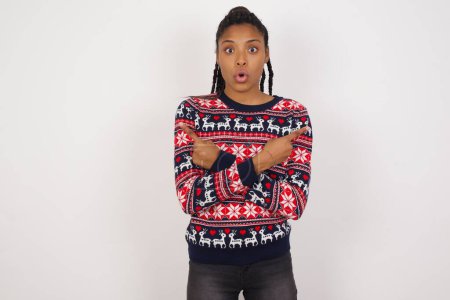 Photo for Confused African American woman wearing Christmas sweater against white wall chooses between two ways, points at both sides with crossed hands, feels doubt. Need your advice. - Royalty Free Image