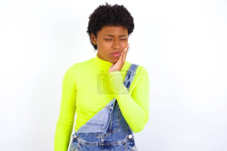 Téléchargez les photos : Jeune femme afro-américaine aux cheveux courts portant globalement du denim contre un mur blanc avec mal de dents - en image libre de droit