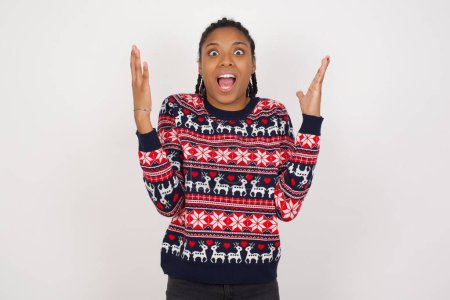 Photo for Joyful excited lucky African American woman wearing Christmas sweater against white wall cheering, celebrating success, screaming yes with clenched fists - Royalty Free Image