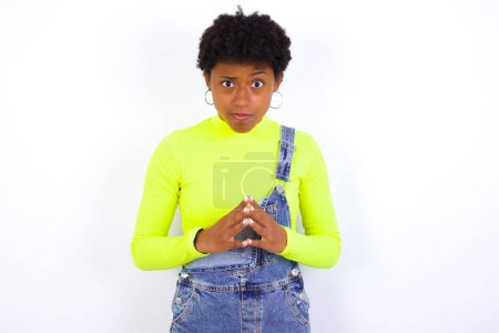 Photo for Young African American woman with short hair wearing denim overall against white wall steepls fingers and looks mysterious aside has great evil plan in mind - Royalty Free Image