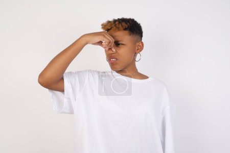 Téléchargez les photos : Mécontent jeune femme afro-américaine portant t-shirt blanc isolé sur fond bouchons nez comme sent quelque chose de puant et désagréable, sent l'aversion. Jeune femme séduisante déteste parfum dégoûtant qui vient de la pièce à côté - en image libre de droit