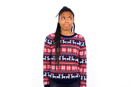 Foto de Sorprendida mujer afroamericana desconcertada vistiendo suéter de Navidad contra la pared blanca curva los labios y tiene mirada preocupada, ve algo horrible en frente. - Imagen libre de derechos