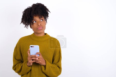 Retrato de mujer joven con aspecto de ensueño, pensando mientras sostiene el teléfono inteligente. Intenta escribir un mensaje.
