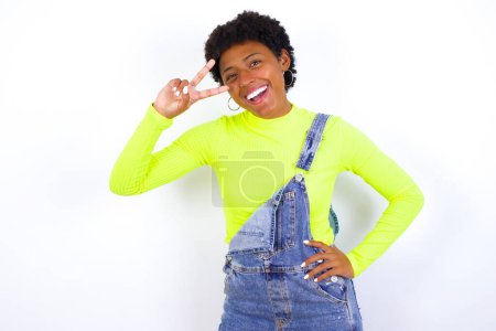 Photo for Young African American woman with short hair wearing denim overall against white wall making v-sign near eyes. Leisure, coquettish, celebration, and flirt concept. - Royalty Free Image