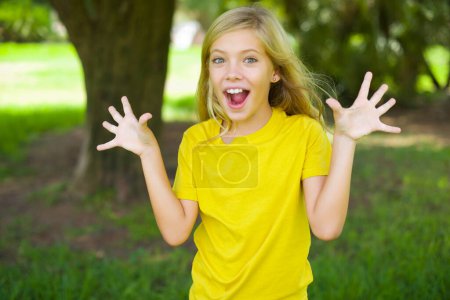 Téléchargez les photos : Délicieuse petite fille caucasienne positive portant un t-shirt jaune debout à l'extérieur ouvre la bouche et les bras paumes vers le haut après avoir obtenu un excellent résultat - en image libre de droit